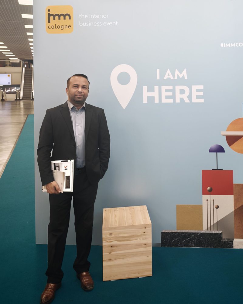 Arslan Azhar Sheikh in a suit beside the imm cologne 2026 'I AM HERE' board